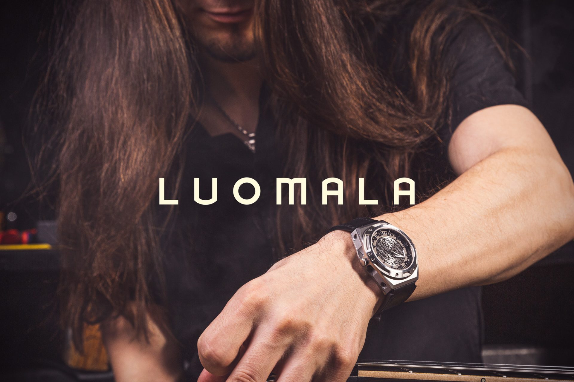 Luomala Watches logo ja taustalla pitkähiuksinen mies Luomala Watches -kello kädessä.