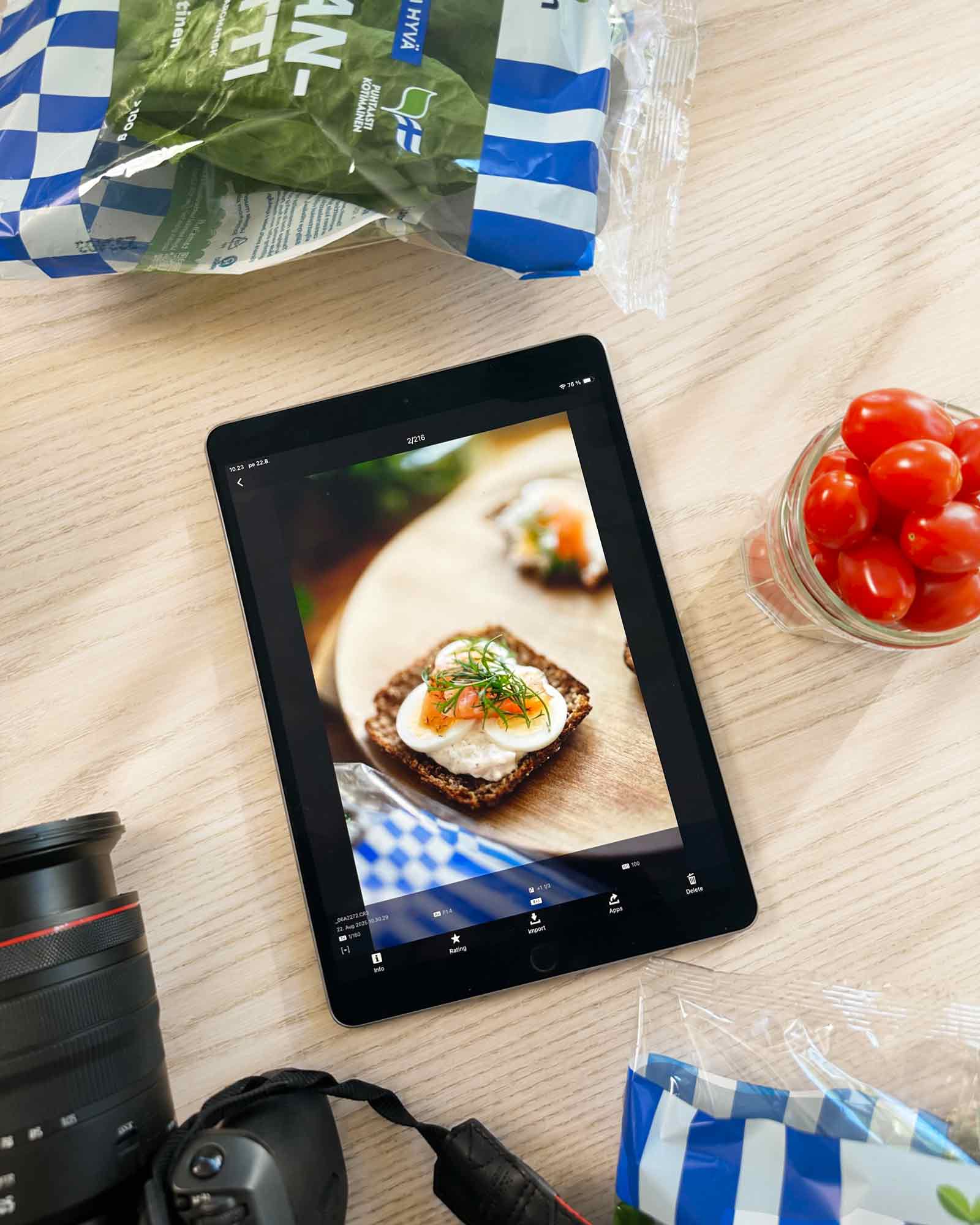 Pöydällä tabletti, jossa näkyy otos leivästä, jonka päällä on tamsin tilliä. Pöydällä ympärillä Tamsin tuotteita ja kamera.